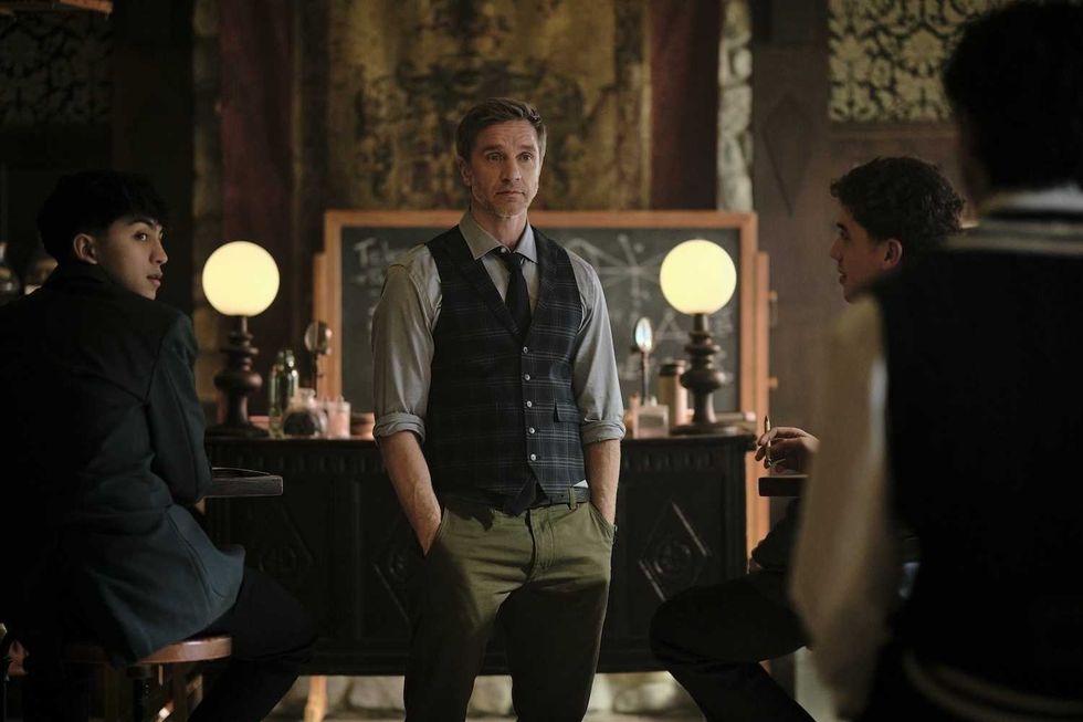 Man in vest stands between three seated people in a dimly lit room with globes and chalkboard.