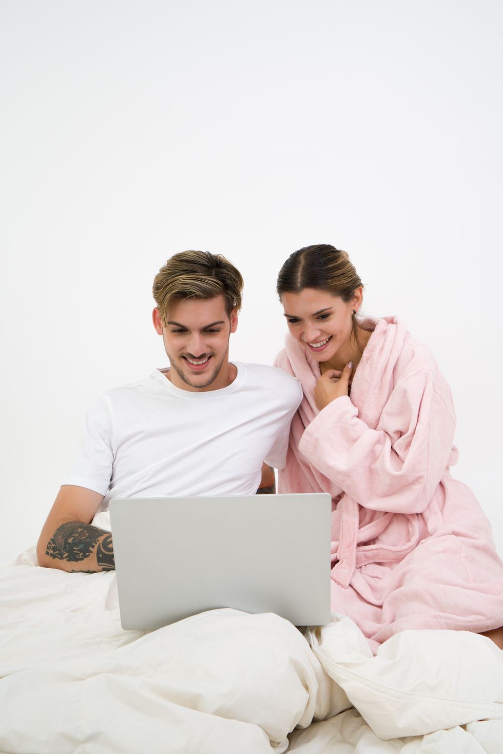 man in white shirt and woman in pink robe looking at a computer screen together in bed