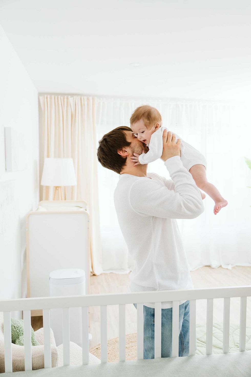Man in white shirt lifts baby in a bright room.