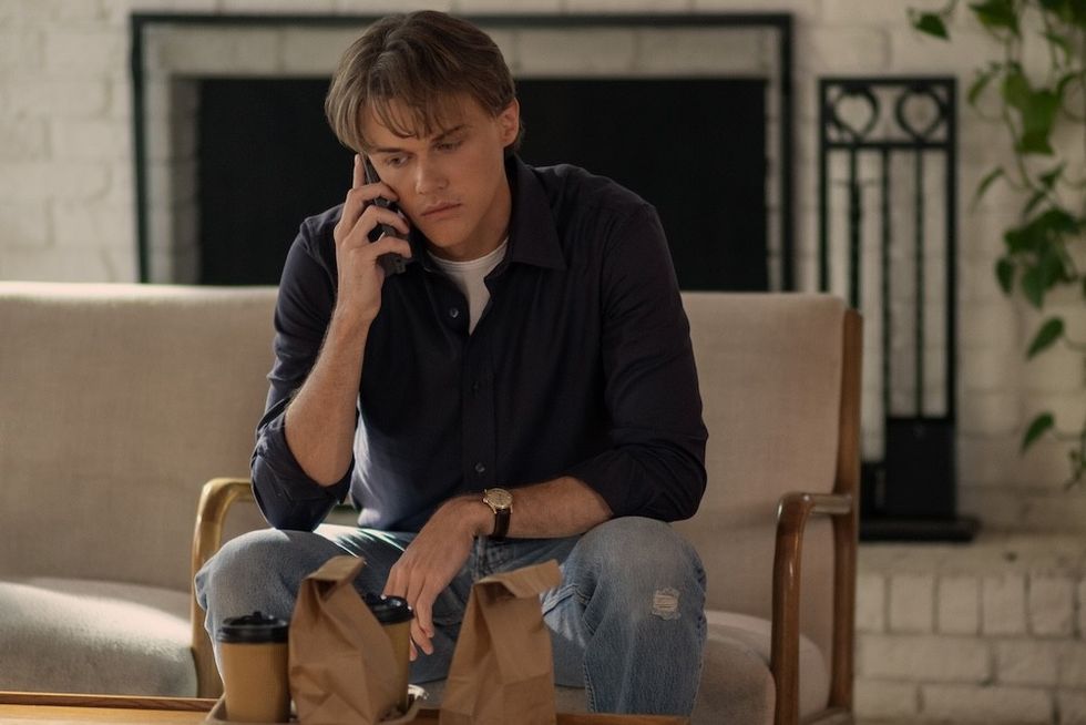 Man sitting on a couch, talking on phone, with takeaway bags on the table chris briney as conrad fisher