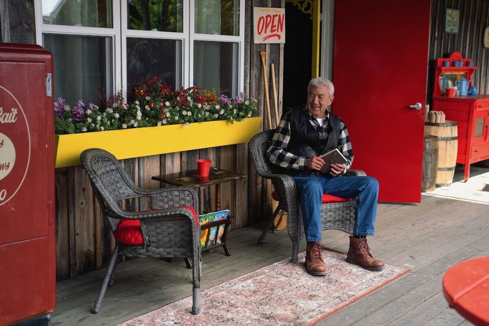 Man sitting with a book outside a rustic shop with a red door and flower boxes.