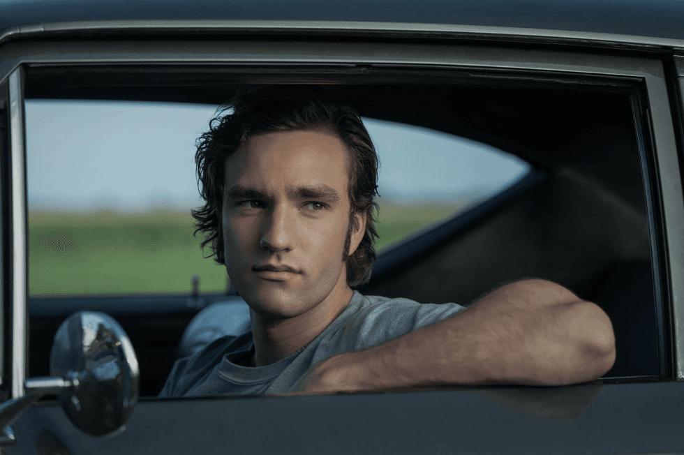 Man with curly hair looking out from a car window against a blurred countryside background.