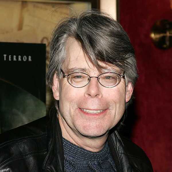 Man with glasses smiling, wearing a black jacket, standing indoors.