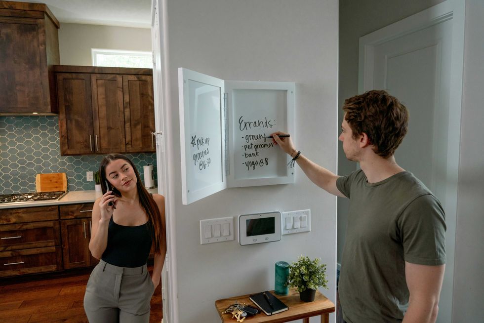 Man writing errands on a whiteboard while a woman talks on the phone in a kitchen.