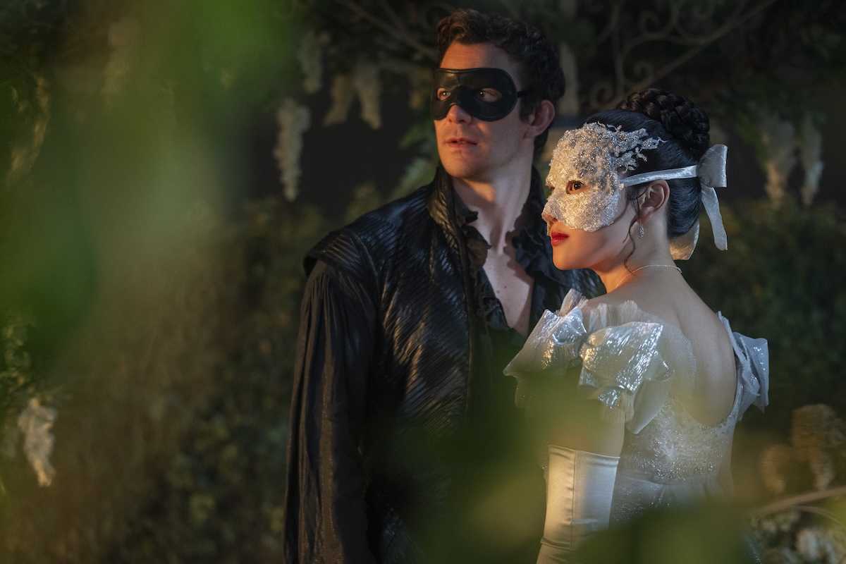 Masked couple in elegant attire in a leafy, dimly lit setting.