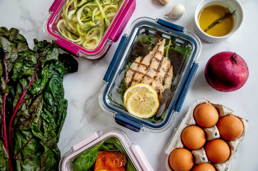 Meal prep containers with grilled chicken, zucchini, salmon, eggs, greens, onion, and garlic.