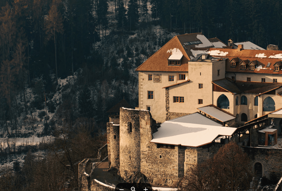 Medieval castle with snow-covered roof, nestled in a mountainous forest landscape.