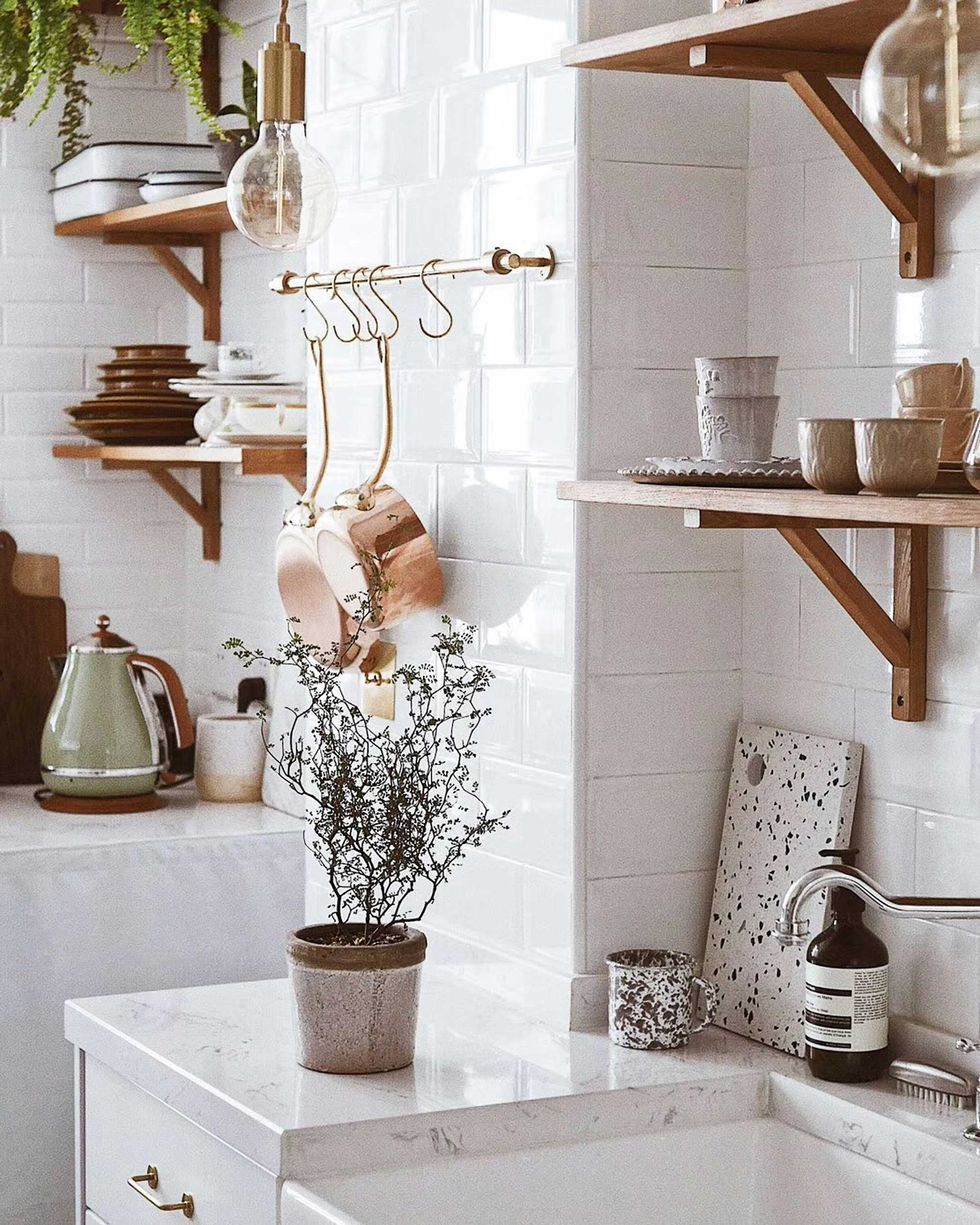Minimal kitchen with white tiles, wooden shelves, hanging copper pans, and potted plant.