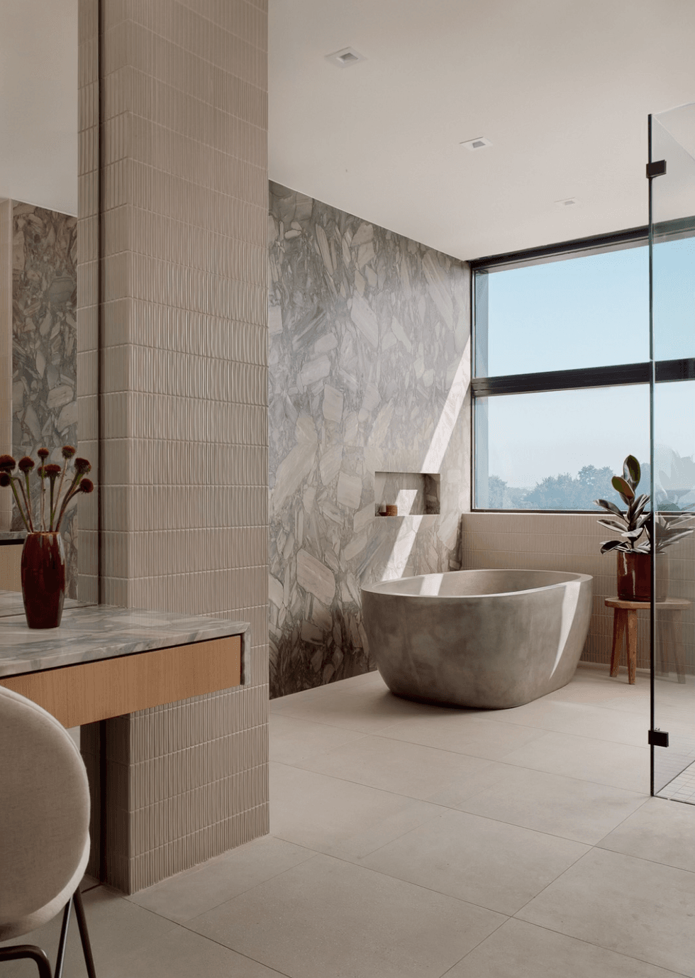 Modern bathroom with marble wall, stone bathtub, large window, and wooden stool with plant.