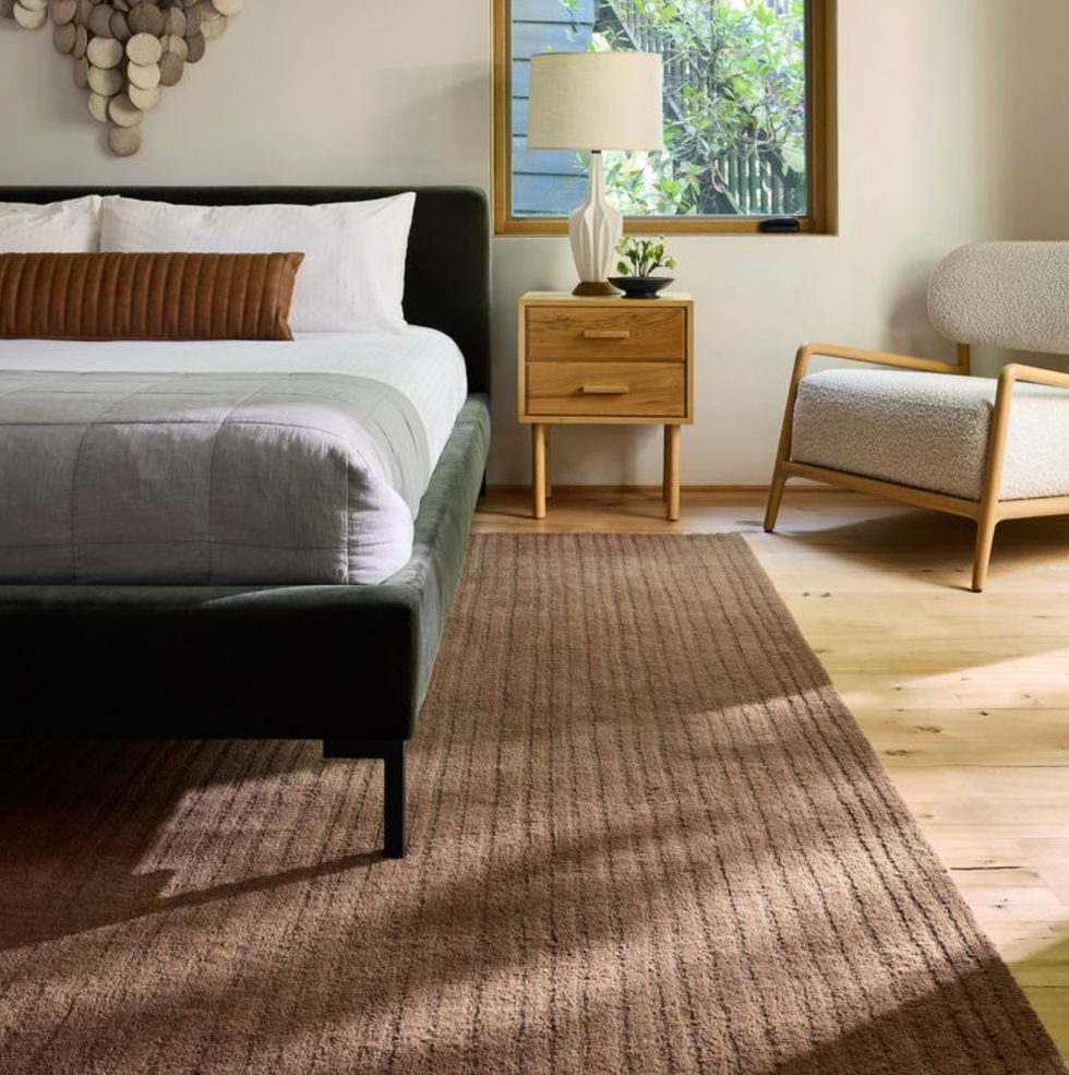 Modern bedroom with a bed, wooden nightstand, lamp, and chair, featuring a large window.