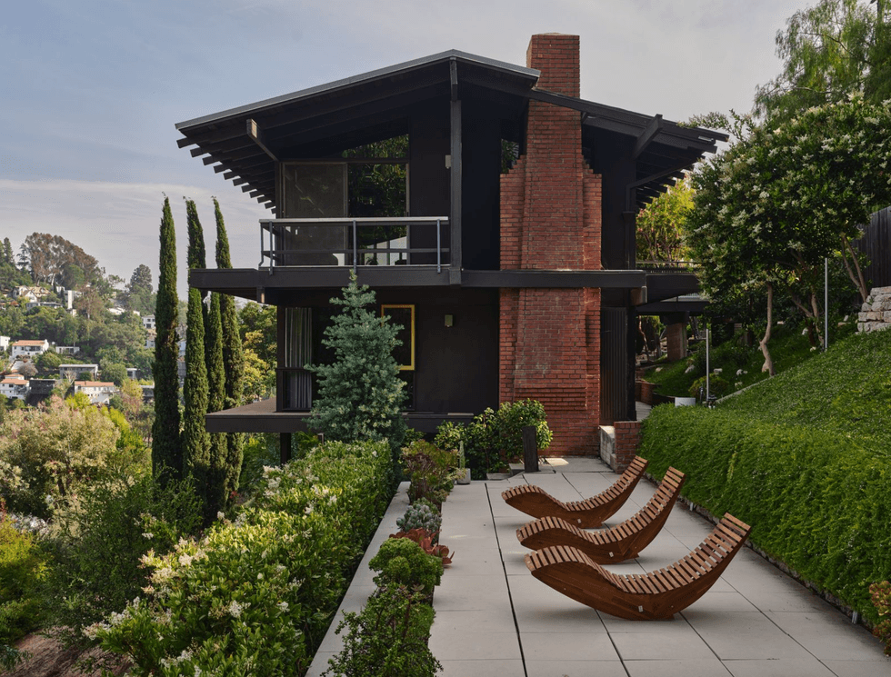 Modern house with brick and wood facade, patio, and wooden lounge chairs surrounded by greenery.