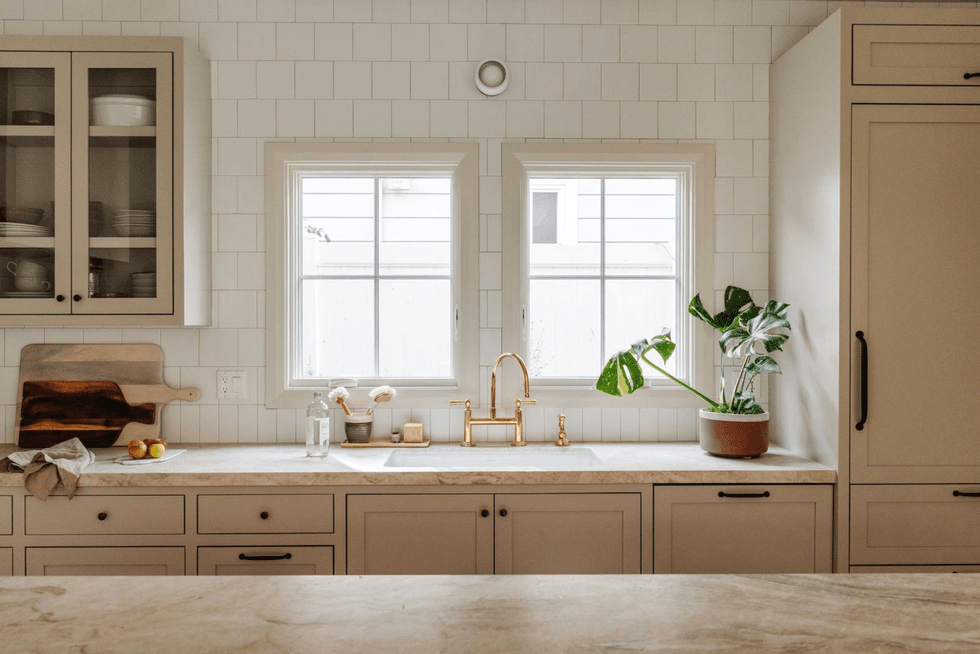 Modern kitchen with beige cabinets, gold faucet, plants, and a large cutting board.