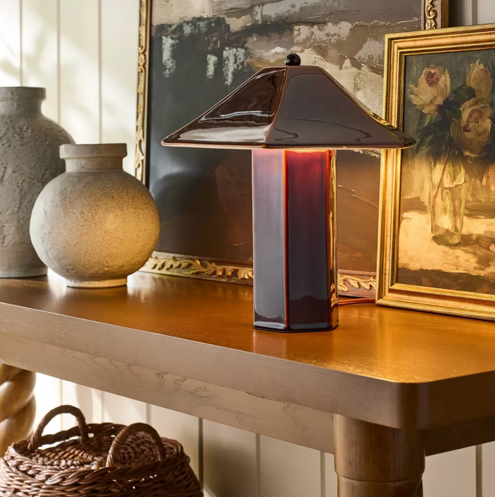 Modern lamp on wooden table with vases, paintings, and a basket in the background.