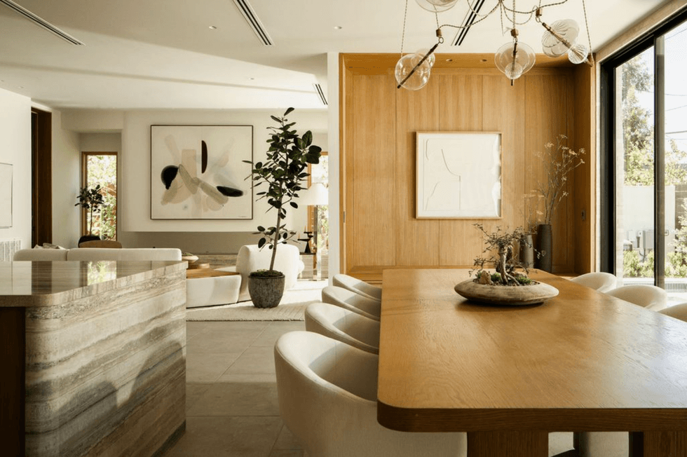 Modern living room with wooden dining table, abstract art, and large windows.