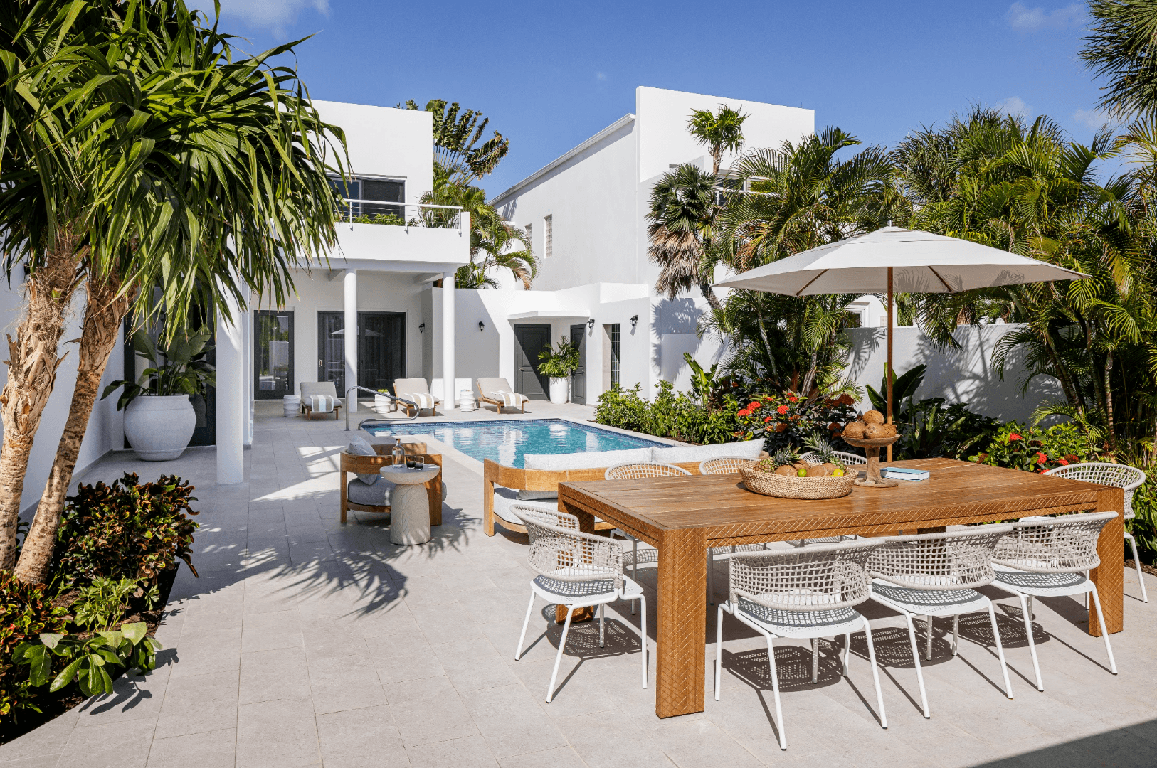 Modern patio with dining set, pool, and lush greenery against a white two-story house.