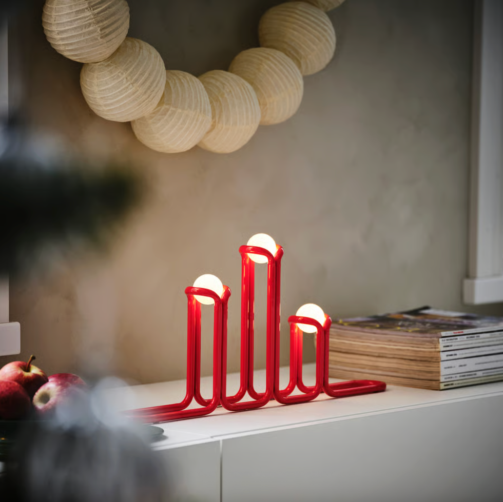 Modern red candle holder with glowing bulbs on a shelf, alongside apples and stacked magazines.