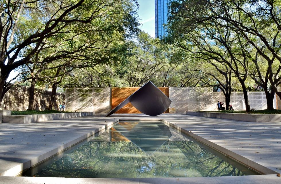Modern sculpture in a park, surrounded by trees, reflected in a rectangular pond.