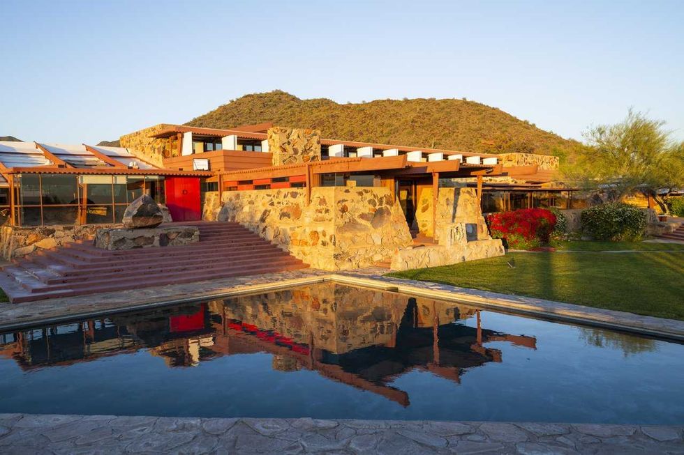 Modern stone building with red accents, reflecting in a tranquil pool, set against a hillside.
