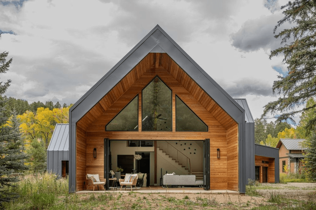 Modern wooden house with large windows, open interior, and outdoor seating amidst trees.