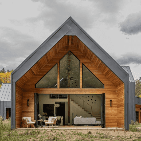Modern wooden house with large windows, open interior, and outdoor seating amidst trees.