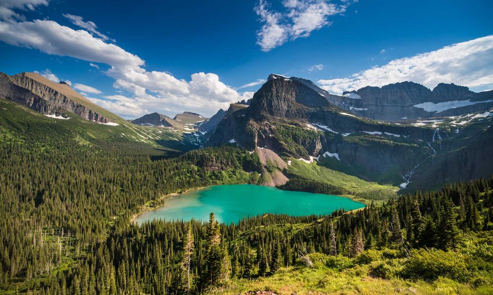 Mountain lake surrounded by dense forest and rugged peaks under a blue sky.