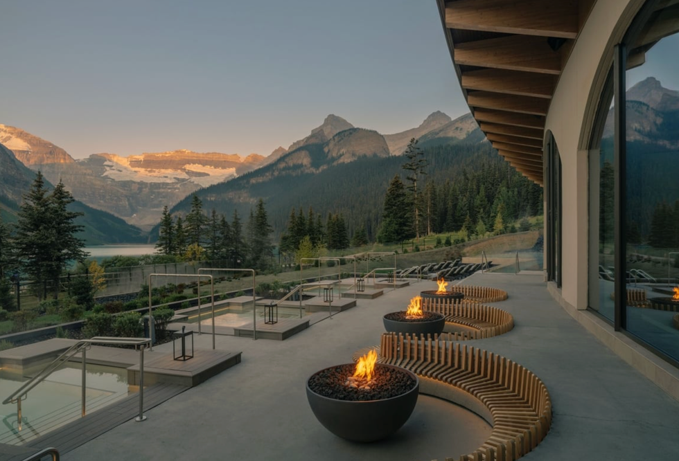 Mountain resort terrace with fire pits, overlooking a serene lake and forested peaks at sunset.