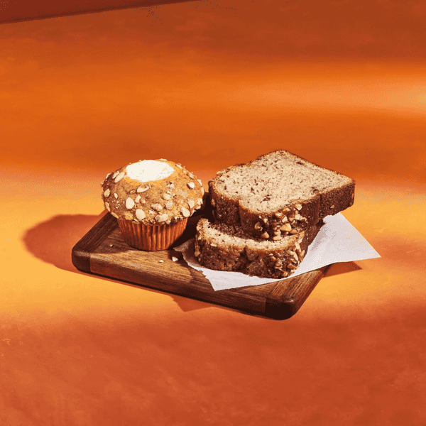 Muffin and sliced banana bread with nuts on a wooden board, on an orange background.