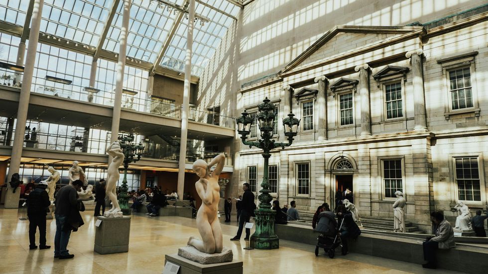 Museum atrium with classical statues, people, and sunlight streaming through glass ceiling.