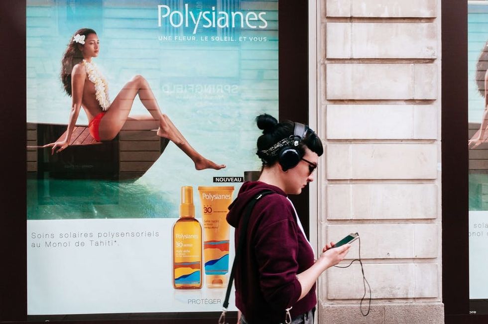 NANTES, LOIRE ATLANTIQUE, BRITTANY, FRANCE - 2016/08/10: Young pretty woman wearing headphones and looking to mobile phone while walking on a street passing by advertising. (Photo by Raquel Maria Carbonell Pagola/LightRocket via Getty Images)
