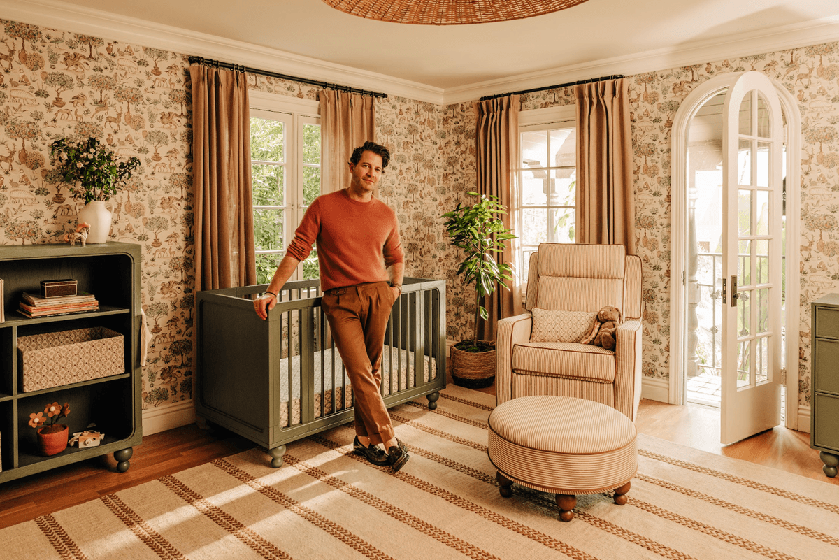 Nate Berkus leaning on crib in a warm, cozy nursery with nature-themed wallpaper and decor.