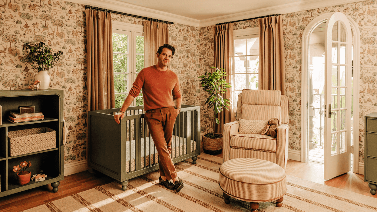 Nate Berkus leaning on crib in a warm, cozy nursery with nature-themed wallpaper and decor.