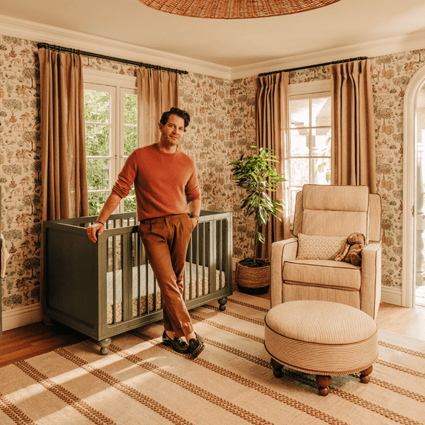Nate Berkus leaning on crib in a warm, cozy nursery with nature-themed wallpaper and decor.