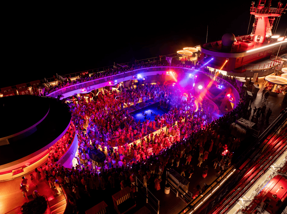Night cruise deck party with vibrant red and purple lighting, crowded with people.