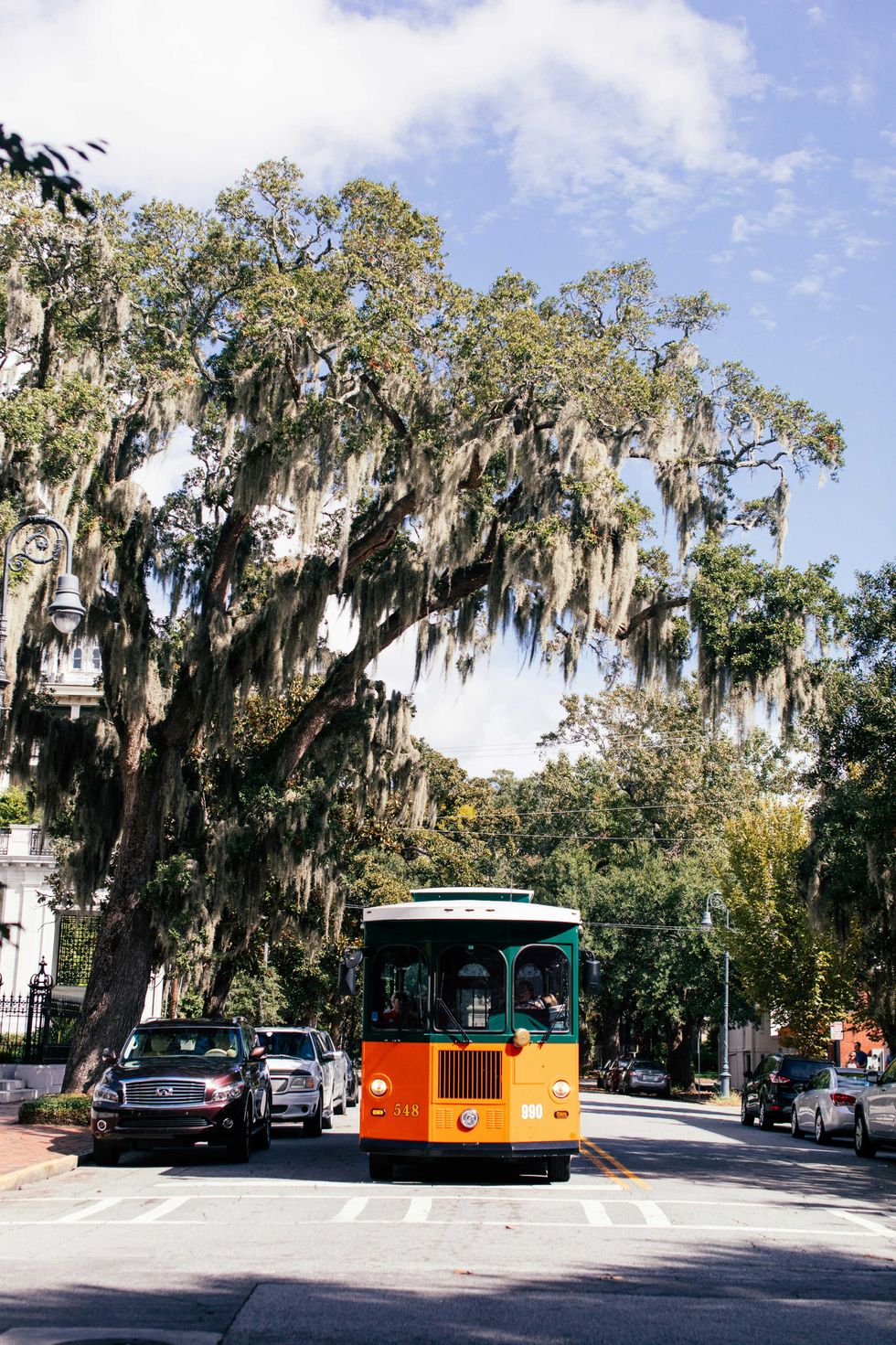 Orange and green trolley under mossy trees on a sunny street.