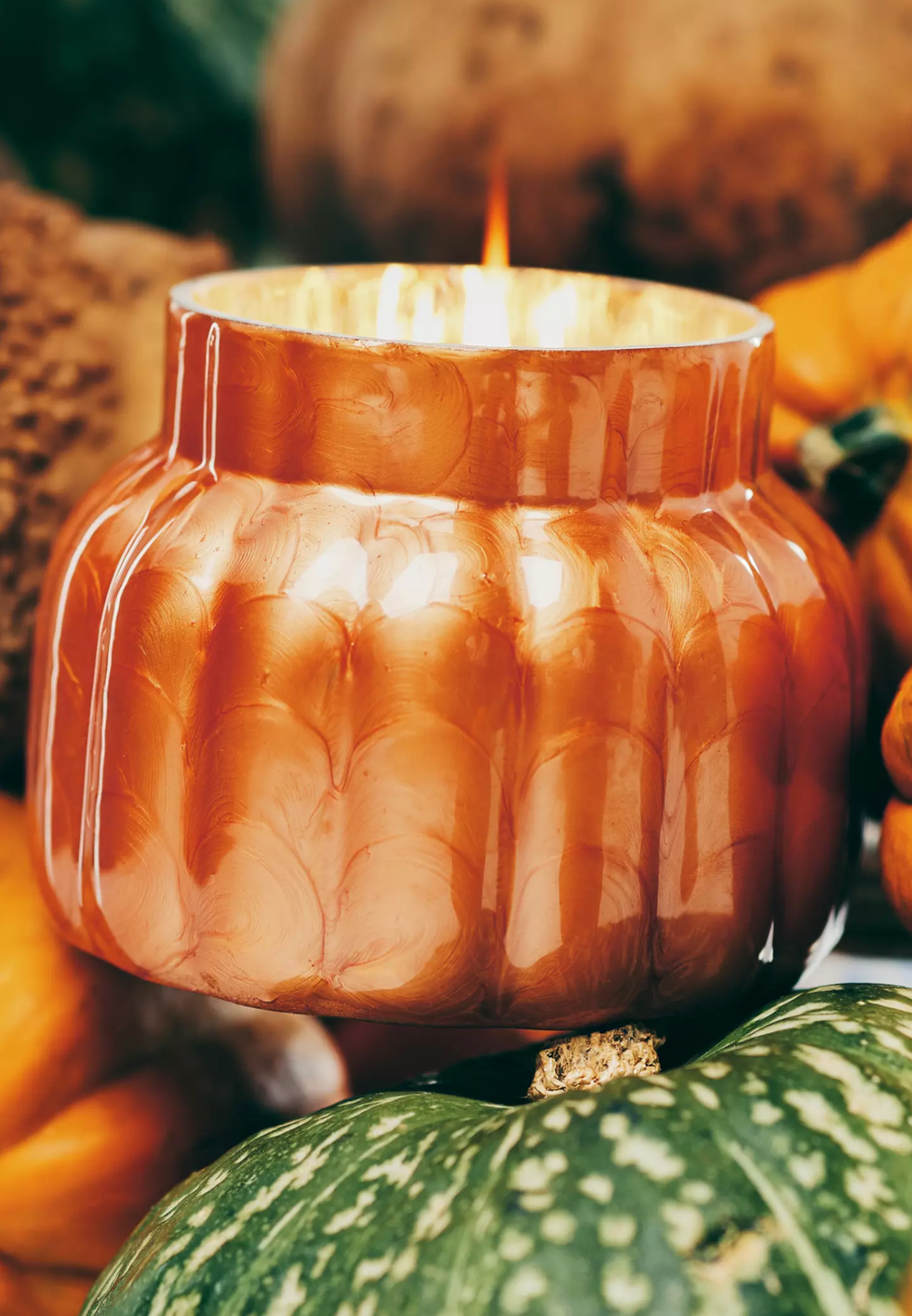 Orange candle with textured glass, surrounded by multicolored pumpkins.
