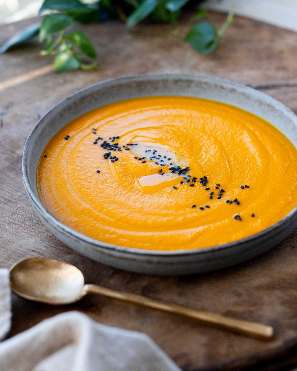 Orange carrot soup in a bowl, garnished with black seeds, with a gold spoon beside it.