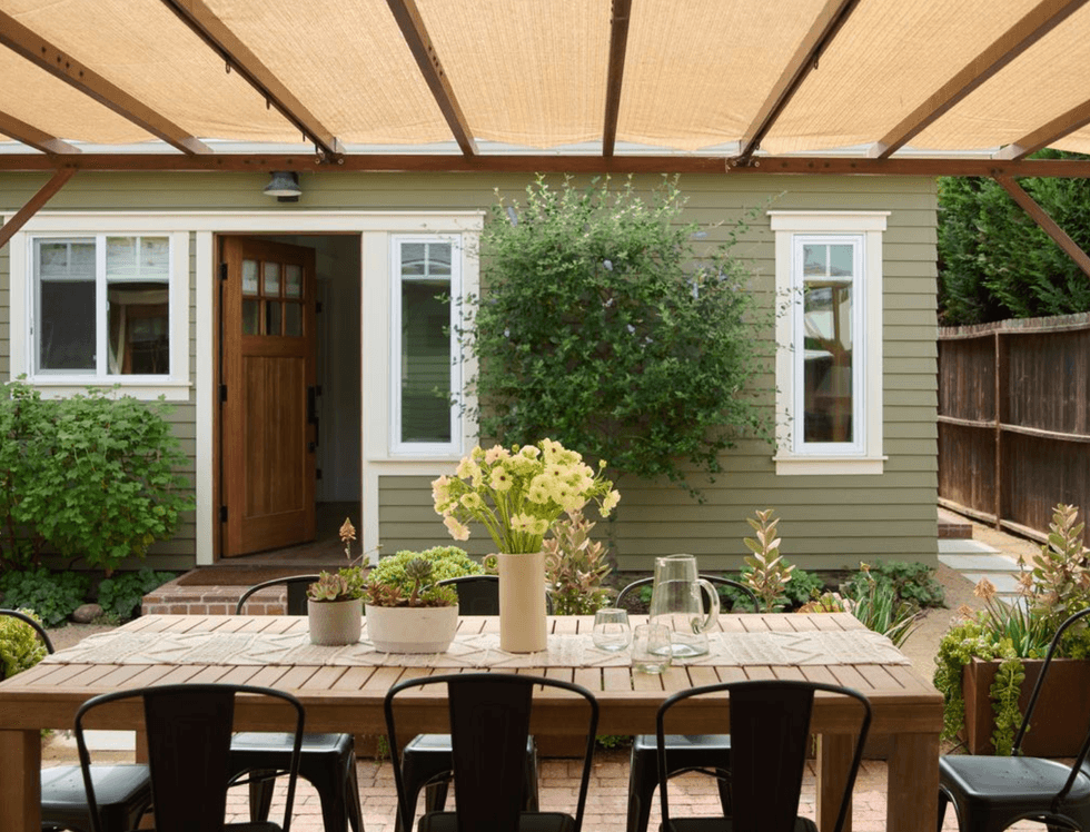 Outdoor dining setup under pergola, facing a green house with open wooden door.