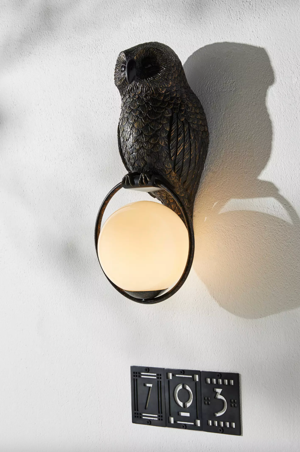 Owl-shaped wall sconce holding a glowing orb, mounted above numbers 7, 0, 3 on a wall.