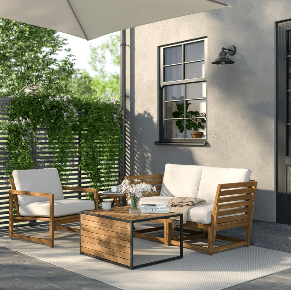 Patio with wooden furniture, a small table, flowers, and greenery under a sunshade.