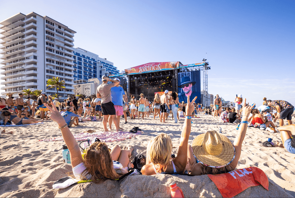 People enjoy a beach concert, lounging on sand with a stage view, under a clear blue sky.