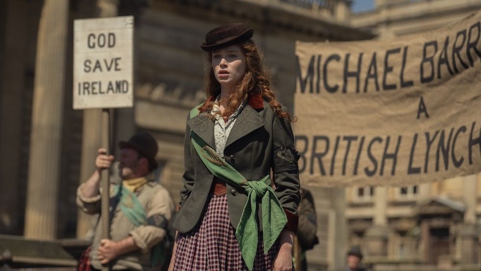 Period drama scene: woman stands with Irish protest signs behind her.
