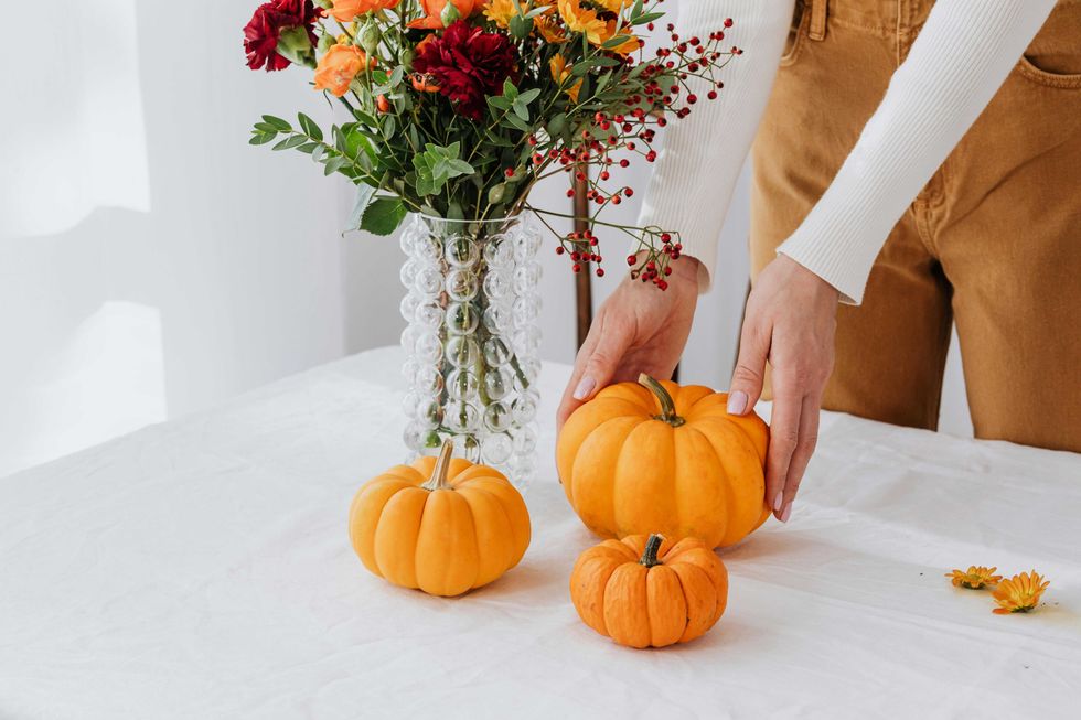 Person arranging small pumpkins on a table with a vase of flowers.