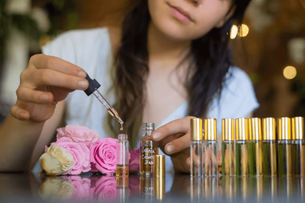 Person blending perfume with dropper, surrounded by roses and bottles.