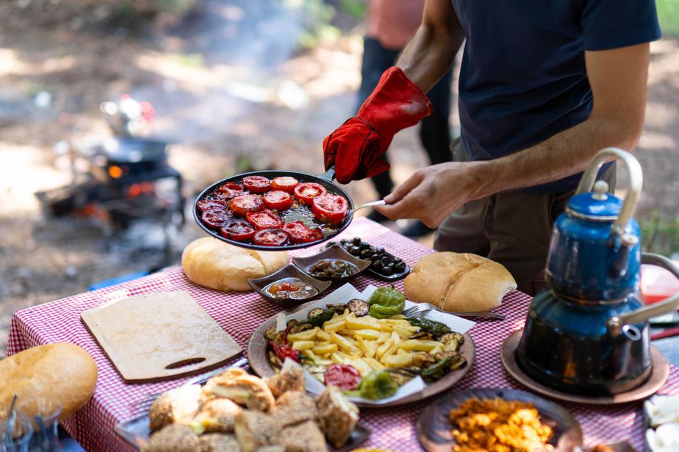 Person cooking outdoors on a table with food, holding a pan of sliced tomatoes.