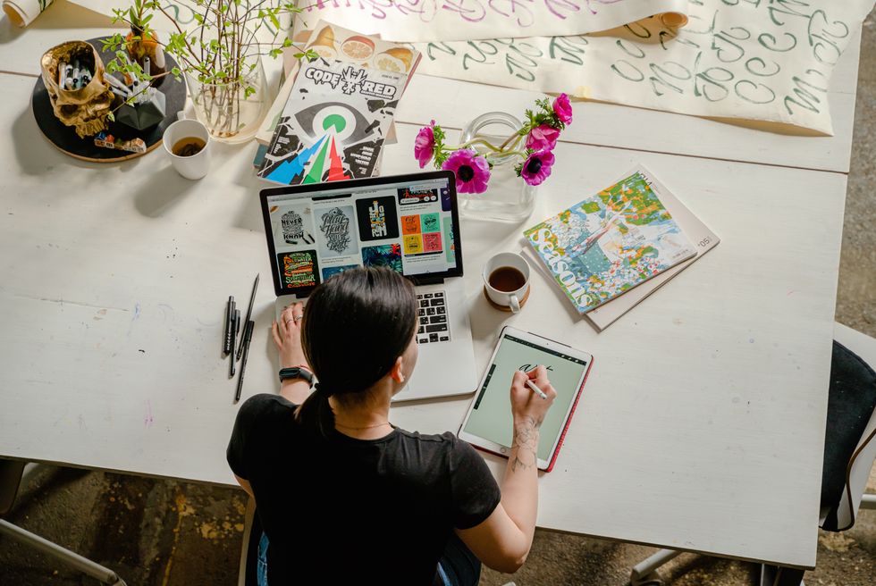 Person designing on tablet, surrounded by flowers and art materials on a table.