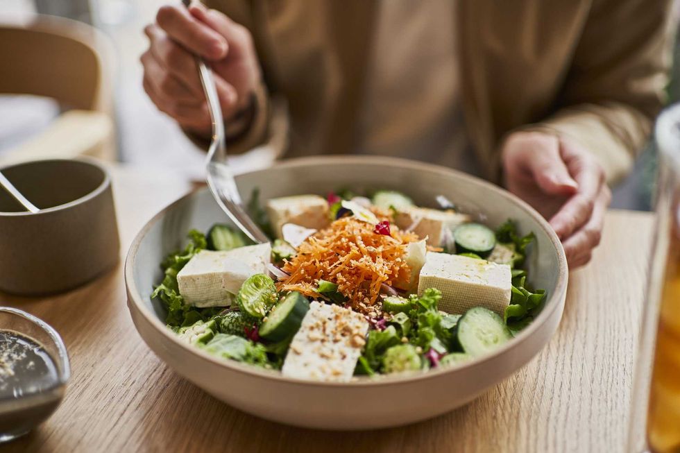 Person eating a fresh salad with tofu, cucumbers, and grated carrots at a wooden table.