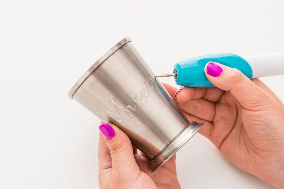 Person engraving "cheers" on a metal cup using an engraving tool.