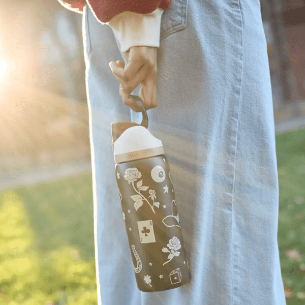 Person holding a decorated bottle by its loop in sunlight, wearing a denim skirt.