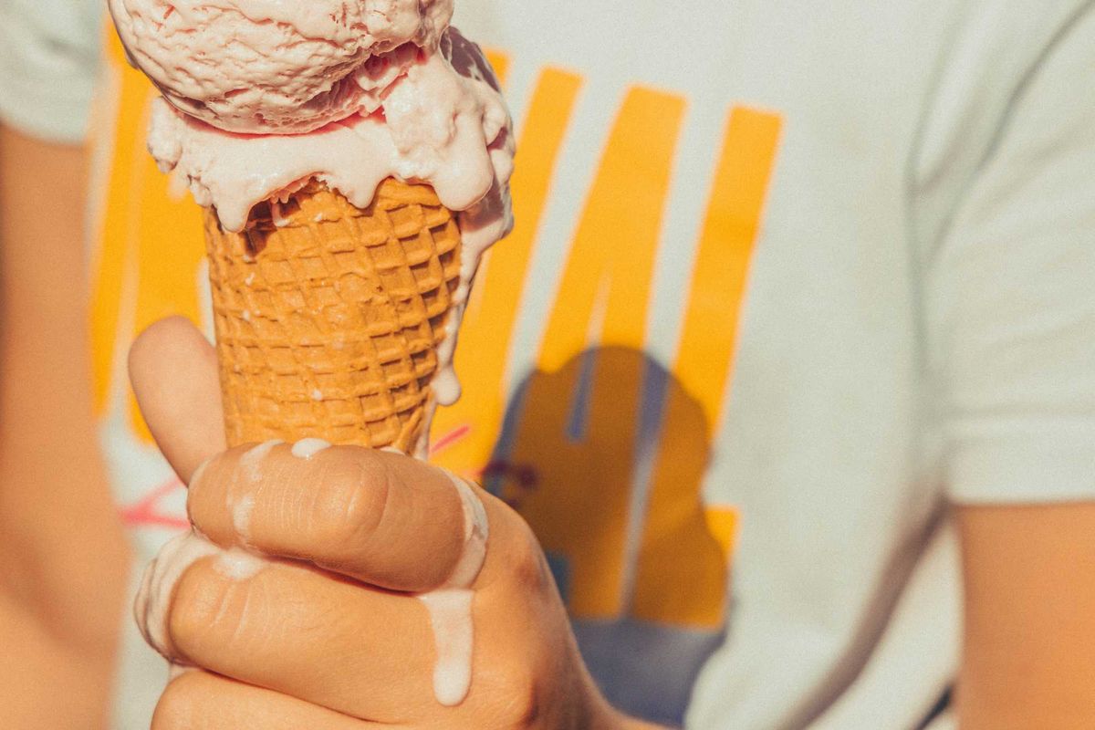 Person holding a melting ice cream cone, wearing a white T-shirt with yellow design.
