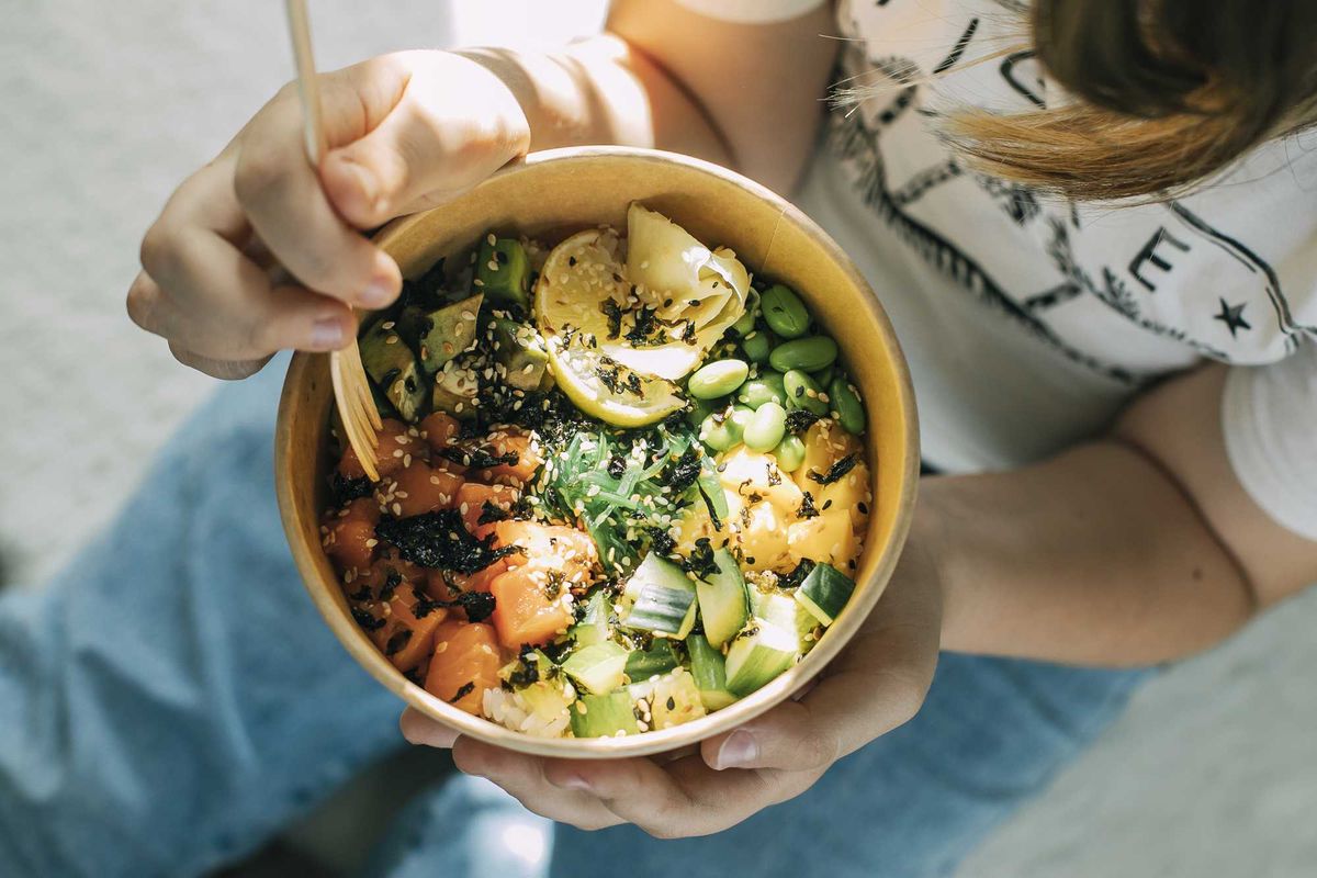 Person holding a poke bowl with salmon, avocado, edamame, and seaweed.